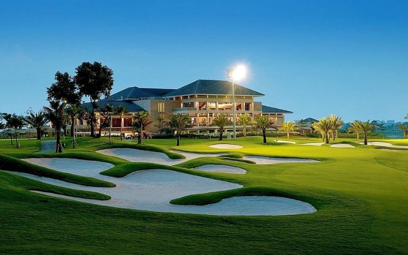 Evening view of Royale Jakarta Golf Club with illuminated clubhouse, manicured fairways, and sculpted bunkers under a clear sky.