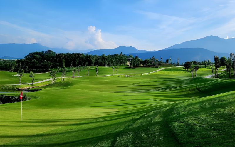 Green fairway at Sapa Grand Golf Club with a flagstick on the left, surrounded by mountains and clear blue skies.