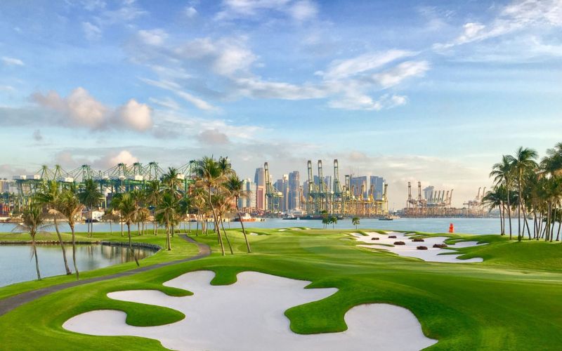 A view of Sentosa Golf Club featuring pristine fairways, sculpted bunkers, and a city skyline rising in the background under a bright sky.