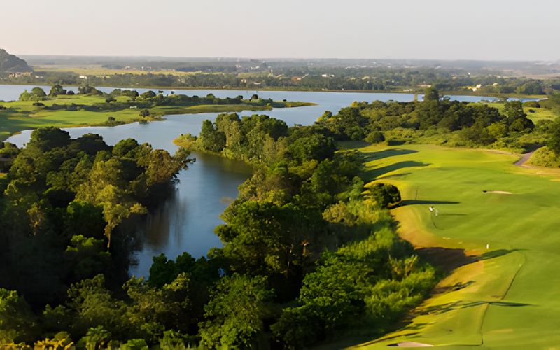 Aerial view of Sky Lake Golf Resort in Hanoi, featuring lush green fairways, a large lake, and surrounding natural landscapes under clear skies.
