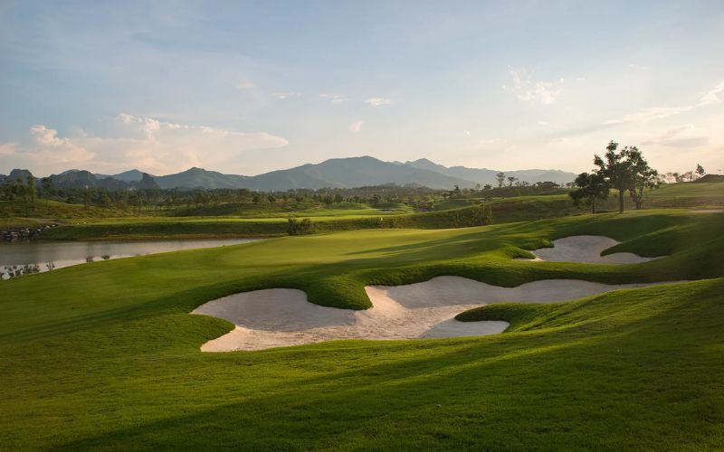 A scenic view of a golf course, featuring a well-maintained green, multiple white sand bunkers, and hills in the background under soft, warm lighting.