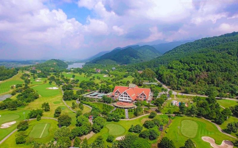 Panoramic view of Tam Dao Golf Resort featuring lush green golf courses, a large building in the center, and surrounding mountains under a partly cloudy sky.