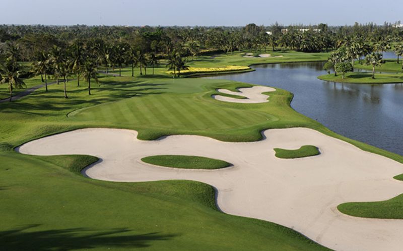 A scenic view of Thai Country Club golf course featuring wide sand bunkers, manicured fairways, and a water hazard under clear skies.