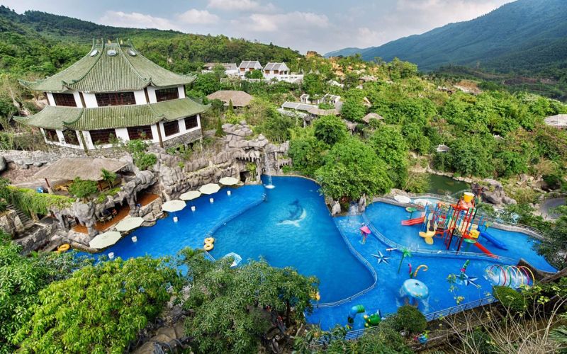 A scenic view of Than Tai Hot Springs Park featuring natural green hills, a large turquoise pool, surrounding lush trees, and a resort-style building nestled in nature.