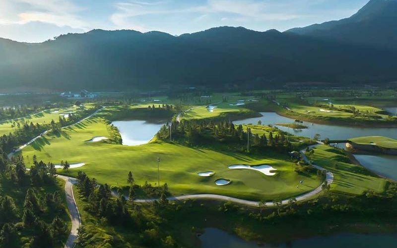 Aerial view of Thanh Lanh Valley Golf Resort, showcasing its lush green fairways, water hazards, and surrounding mountains under soft sunlight.
