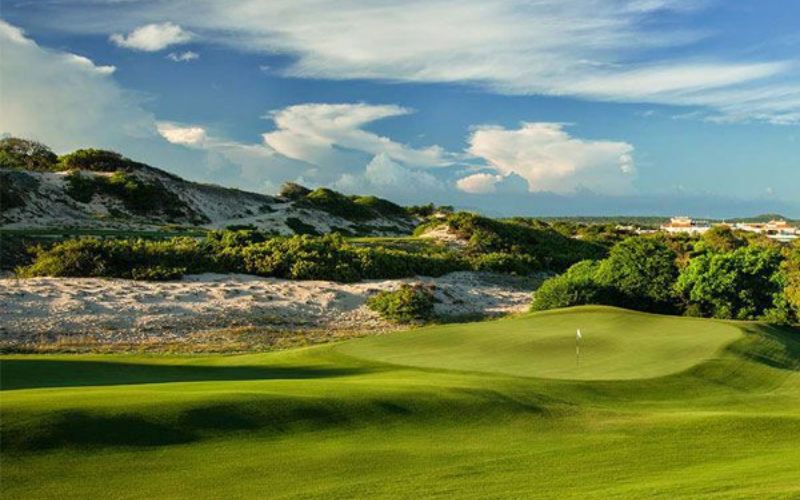 The Bluffs Ho Tram Strip golf course in Vietnam, showing a green with surrounding white sand dunes and blue sky with clouds.