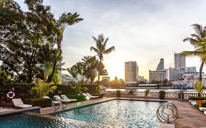 Riverside swimming pool at The Peninsula Bangkok with city skyline and palm trees at sunset.