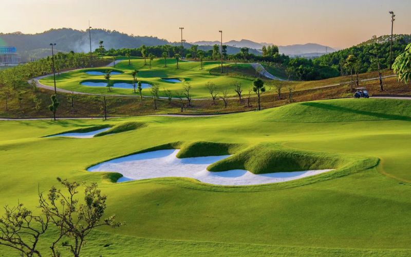 The Van Lang Empire Golf Club course showing a lush, rolling green landscape, prominent sand bunkers, and distant hills at sunset.