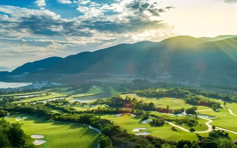 Sunlit Vinpearl Phu Quoc golf course surrounded by lush greenery and mountain views under a partly cloudy sky.
