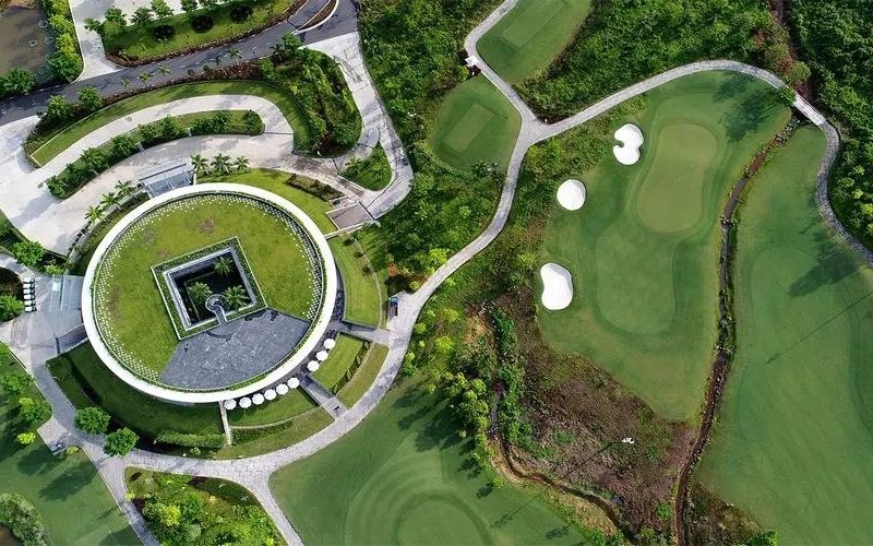 Aerial view of Ba Na Hills Golf Club showing a circular clubhouse surrounded by lush greenery, curved pathways, and a section of the landscaped golf course with fairways and bunkers.