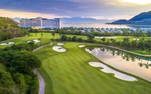 A panoramic view of Vinpearl Golf Nha Trang featuring lush fairways, sand bunkers, water hazards, and mountains in the background under a soft sky.