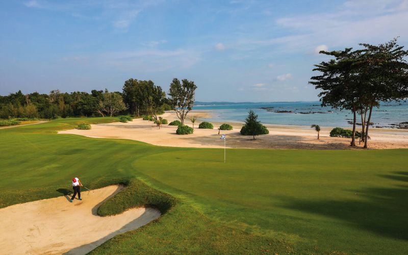 The Els Club Malaysia golf course featuring a golfer in a greenside bunker, manicured fairway, sandy coastline, and ocean views.