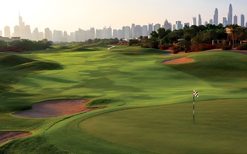 Montgomerie Golf Club Dubai in the evening, showcasing wide fairways, rolling greens, water features, and a distant city skyline at sunset.