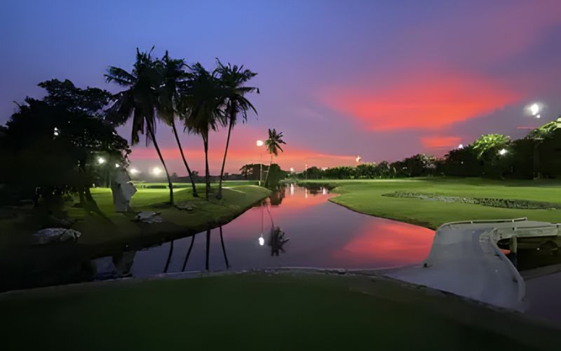 A vibrant sunset over the illuminated fairways and water features at Summit Windmill Golf Club in Thailand, with palm trees reflected on the calm surface.