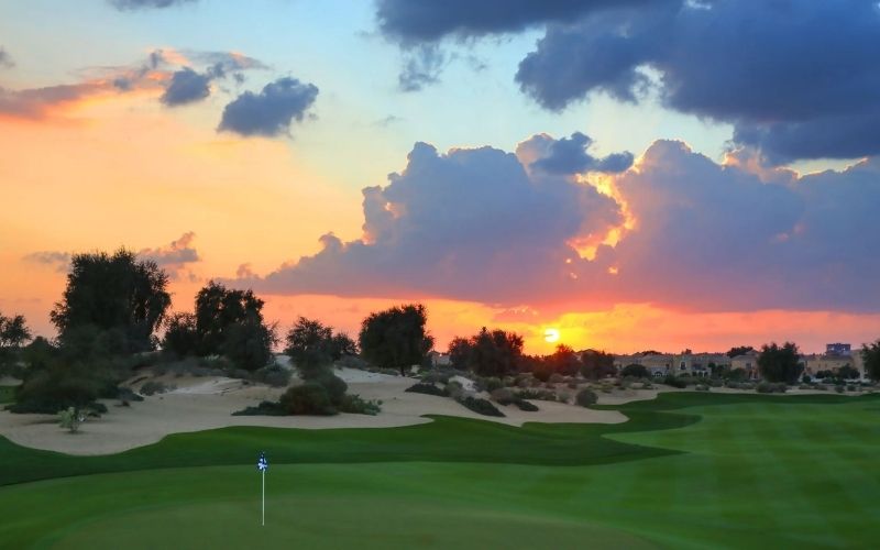 View of The Els Club Dubai golf course at sunset, with illuminated fairways, palm trees, and a dramatic sky over the desert landscape.