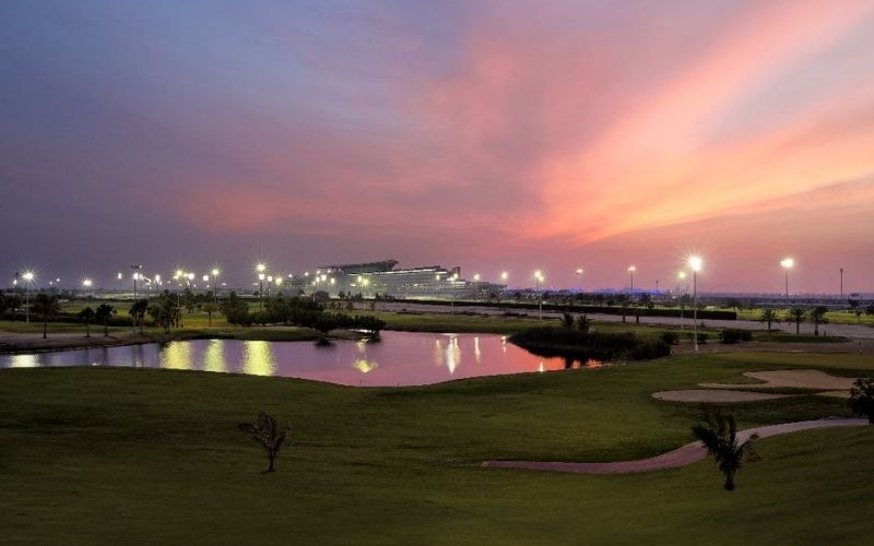 Evening view of The Track Meydan Golf course in Dubai, with illuminated greens, water features, and the city skyline glowing under a colorful sunset sky.