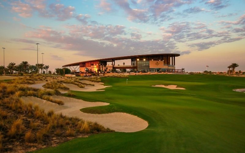 Trump International Golf Club Dubai at dusk, featuring manicured fairways, sand bunkers, and the clubhouse glowing under warm evening light.
