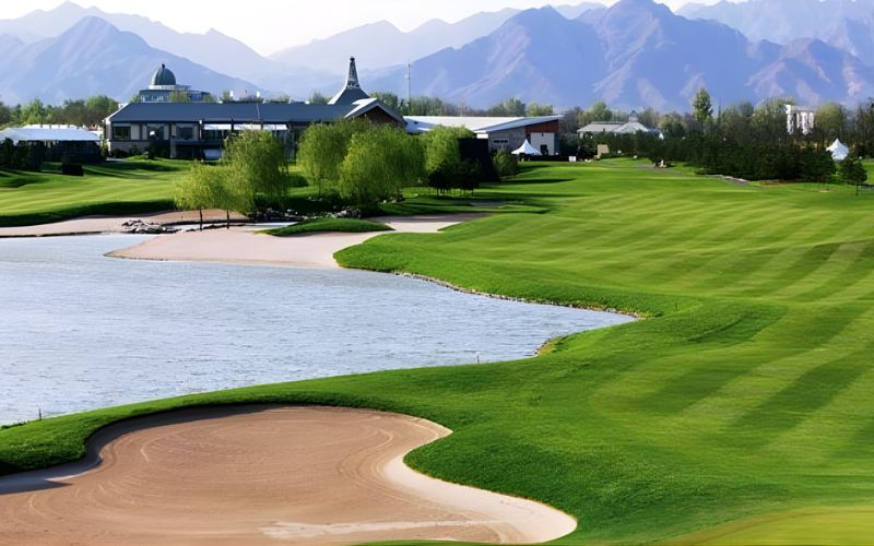 Grand Dynasty Golf Club fairway running alongside a lake with a sand bunker in the foreground and mountains in the background under clear skies.